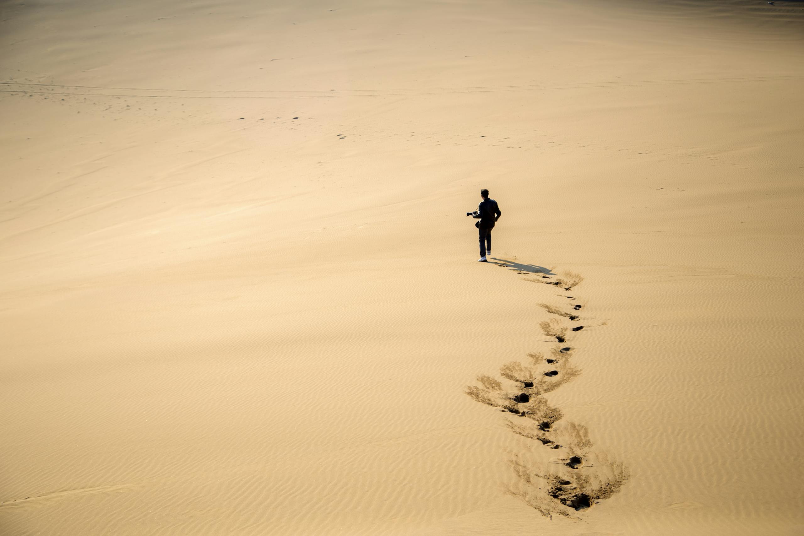 A photographer walking across vast sand dunes in the Iranian desert, leaving footprints behind.