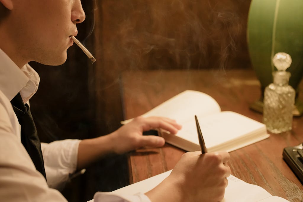 Asian man writing at a desk, cigarette in mouth, warm indoor setting.